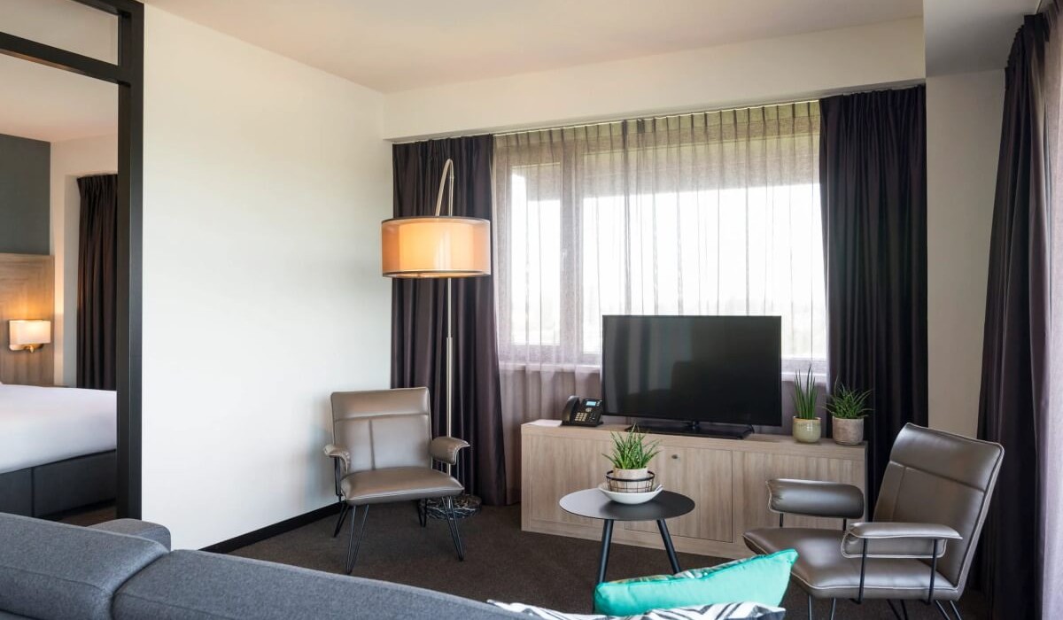 Adagio Amsterdam City South - Il salotto del Junior Apartment offre un angolo TV con canali satellitari, un comodo divano e due poltrone, creando un ambiente accogliente ideale per momenti di relax.