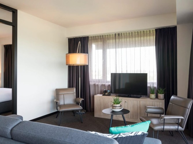Adagio Amsterdam City South - Il salotto del Junior Apartment offre un angolo TV con canali satellitari, un comodo divano e due poltrone, creando un ambiente accogliente ideale per momenti di relax.
