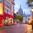 Al mattino, Place du Tertre a Montmartre si anima tra tavolini di caffè e artisti che preparano le loro tele, con la Basilica del Sacro Cuore che domina lo sfondo parigino.