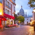 Al mattino, Place du Tertre a Montmartre si anima tra tavolini di caffè e artisti che preparano le loro tele, con la Basilica del Sacro Cuore che domina lo sfondo parigino.
