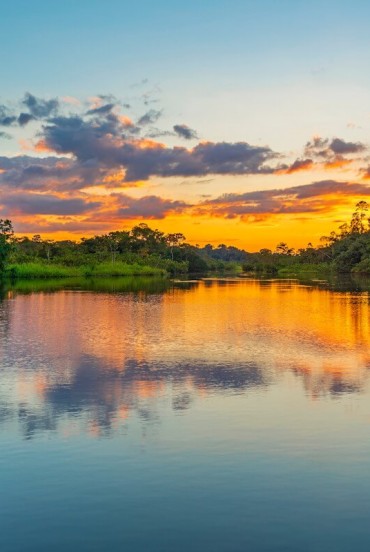 Il sole cala sulle rive di un fiume amazzonico in Ecuador, tingendo l’acqua di riflessi dorati e la foresta di silenzi magici, tra canti lontani e natura incontaminata.