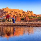 Il villaggio fortificato di Ait Benhaddou, patrimonio UNESCO del Marocco, si riflette nell’acqua tra cammelli, mura in terra e luce del tramonto.