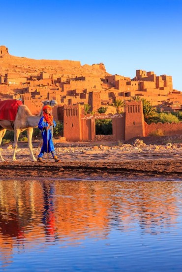 Il villaggio fortificato di Ait Benhaddou, patrimonio UNESCO del Marocco, si riflette nell’acqua tra cammelli, mura in terra e luce del tramonto.