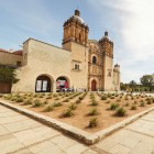 La Chiesa e il Convento di Santo Domingo a Oaxaca risplendono sotto un cielo limpido, affacciati su un giardino di agavi che incornicia l’elegante facciata barocca in una scena luminosa e vivace.