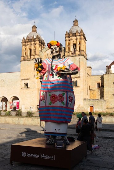 Catrina colorata davanti alla Chiesa di Santo Domingo a Oaxaca: la figura del Día de los Muertos spicca tra l’architettura barocca mentre i visitatori passeggiano nella piazza.