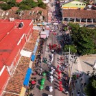 Sfilata colorata a Tehuantepec vista dall’alto: gruppi in abiti tradizionali sfilano con ombrelli rossi, bianchi e verdi, mentre la folla assiste lungo il corso tra edifici coloniali