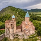 Veduta aerea della fortezza di Gremi in Kakheti: torri in pietra e chiesa con tetti in cotto dominano la collina, tra verdi vallate e montagne della Georgia orientale.