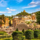 Nel cuore di Tbilisi, le cupole delle terme sulfuree emergono tra giardini curati e case colorate, con la collina e una chiesa che dominano il panorama.
