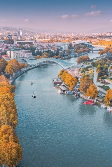 Vista aerea di Tbilisi, con il fiume Mtkvari (Kura) che attraversa la città, il Ponte della Pace e i parchi lungo il lungofiume tra architettura storica e moderna.