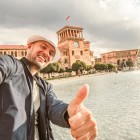 Turista sorridente scatta un selfie davanti alle fontane di Piazza della Repubblica a Yerevan. Sullo sfondo la torre dell’orologio del Palazzo del Governo e l’elegante architettura armena.