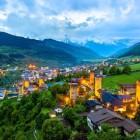 Veduta di Mestia al crepuscolo, con torri medievali illuminate tra verdi vallate e case tradizionali, incorniciata dalle maestose montagne del Caucaso.