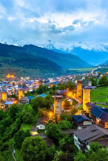 Veduta di Mestia al crepuscolo, con torri medievali illuminate tra verdi vallate e case tradizionali, incorniciata dalle maestose montagne del Caucaso.