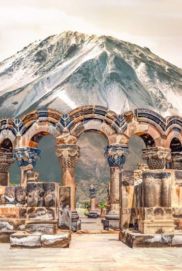 Antiche rovine della Cattedrale di Zvartnots in Armenia, con eleganti archi in pietra scolpita sullo sfondo del maestoso Monte Ararat innevato.