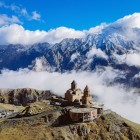 Veduta spettacolare della Chiesa della Trinità di Gergeti, arroccata tra le montagne del Caucaso con il massiccio del Monte Kazbek sullo sfondo e nuvole che avvolgono la valle.
