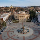 Veduta aerea di Kutaisi con la piazza centrale e la fontana Colchide, circondata da edifici storici. Sullo sfondo la Cattedrale di Bagrati domina dolcemente la città.