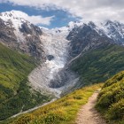 Sentiero panoramico tra prati verdi e luce di montagna, ai piedi del ghiacciaio dello Shkhara. Il percorso accompagna lo sguardo verso le vette del Caucaso, tra silenzio e spazi aperti.