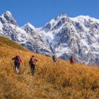 Trekking tra erbe dorate ai piedi del ghiacciaio del monte Shkhara nello Svaneti. Il sentiero sale tra silenzi e panorami aperti, mentre le vette innevate accompagnano ogni passo.