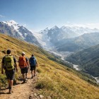 Tre escursionisti percorrono il sentiero del passo Chkhunderi, tra prati dorati e silenzi d’alta quota. Davanti, la valle si apre verso il fiume e le vette innevate del Caucaso.