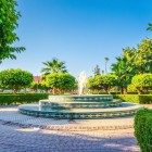 Fontana centrale tra vialetti curati, palme e alberi rigogliosi. Sullo sfondo la Moschea Koutoubia di Marrakesh, tra geometrie armoniose e luce intensa.