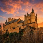 Il profilo fiabesco dell'Alcázar di Segovia domina lo sperone roccioso mentre il cielo al tramonto avvolge torri, merli e mura in una luce dorata che esalta l'eleganza dell'architettura medievale.