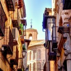 Calle de San Bartolomé svela la Toledo più autentica: balconi in ferro, dimore storiche e l’abside mudéjar della chiesa medievale, accanto al Palazzo Pantoja y Angulo.