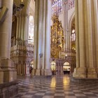 Possenti pilastri gotici scandiscono le navate della Cattedrale di Toledo; il pavimento geometrico conduce lo sguardo verso il retablo dorato, incorniciato da archi e vetrate.