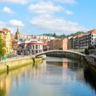 La Ría di Bilbao attraversa il Casco Viejo tra ponti, passeggiate alberate e facciate colorate, conducendo lo sguardo verso il Mercado de la Ribera, cuore della vita cittadina.