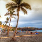 Sulla Playa de La Arena, a Puerto de Santiago, palme e ombrelloni accompagnano la spiaggia di sabbia vulcanica nera, affacciata sull’Atlantico sotto un suggestivo cielo al tramonto.