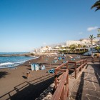 Il lungomare di Playa de La Arena, a Puerto de Santiago, costeggia la caratteristica spiaggia di sabbia nera, tra palme, terrazze panoramiche e il blu dell’Atlantico.