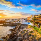Al tramonto, il lungomare di Puerto de Santiago segue la costa vulcanica tra palme, terrazze e piscine affacciate sull’Atlantico, mentre il cielo si accende di calde sfumature dorate.