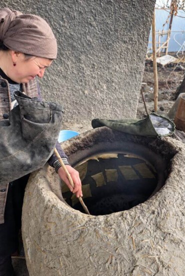 La padrona di casa prepara il pranzo nel grande kazan inserito nel focolare in terra, mostrando una tecnica tradizionale ancora presente nella cucina familiare del villaggio.